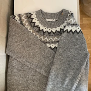 Grå stickad tröja med mönster - Säljer en grå stickad tröja från 157 med klassiskt nordiskt mönster i vitt och mörkgrått runt halsen. Tröjan har rund halsringning och långa ärmar med ribbade muddar. Perfekt för kyliga dagar!