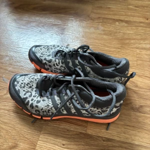 Adidas träningsskor med leopardmönster - Säljer ett par Adidas träningsskor med grått och svart leopardmönster, orange detaljer och svarta snören. Skorna har låg profil, platt sula och är perfekta för dig som vill sticka ut på gymmet eller löprundan, i mycket bra skick! 