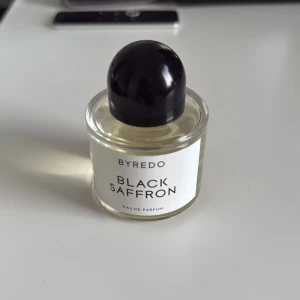Byredo Black Saffron Eau de Parfum - Byredo Black Saffron Eau de Parfum i en rund, genomskinlig glasflaska med svart rund kork. Parfymen har en ljusgul vätska och en stilren vit etikett med svart text. En exklusiv och modern doft från det populära märket Byredo. Ca 40ml kvar, har kvitto från nk