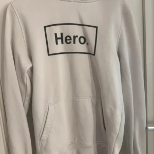 Vit hoodie med tryck från Hero. - Säljer en vit hoodie med svart tryck 'Hero.' på bröstet. Tröjan har huva och magficka, samt långa ärmar. Perfekt för en avslappnad och stilren look.