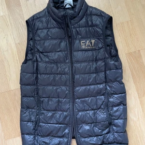 Svart dunväst från Emporio Armani EA7  - Snygg svart dunväst från Emporio Armani EA7 med guldfärgad logga på bröstet. Västen har quiltad design och dragkedja framtill. Nypris 1800 kr, men säljer för 700, pris kan diskuteras