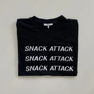 Svart t-shirt med text från GANNI - Svart t-shirt från GANNI med vit text 'SNACK ATTACK' tryckt tre gånger på framsidan. Klassisk rund hals och korta ärmar. Perfekt för en avslappnad och cool stil.