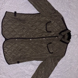 Olivgrön quiltad jacka från Zara Man - Säljer en olivgrön quiltad jacka från Zara Man med svarta detaljer. Jackan har knappar och dragkedja framtill, två fickor med lock och lång ärm. Perfekt för övergångssäsonger 