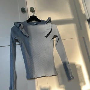 Ljusgrå ribbad tröja med volang från Zara - Supersnygg ljusgrå ribbad tröja från Zara med långa ärmar och volangdetaljer på axlarna. Perfekt för dig som vill ha något stilrent men ändå med en twist. Sitter tajt och är riktigt bekväm.