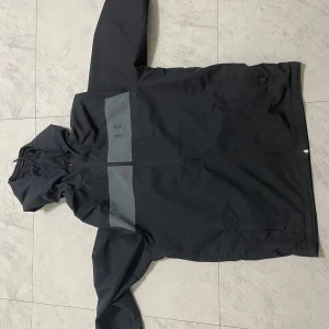 Svart vindjacka från Under Armour - Svart vindjacka med grått parti över bröstet och Under Armour-logga. Jackan har huva, dragkedja framtill och två sidofickor. Perfekt för blåsiga dagar och enkel att matcha med det mesta.