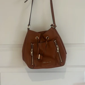 Brun axelväska från Karen Millen - Säljer en snygg brun axelväska från Karen Millen i skinn med två dragkedjor framtill och justerbar axelrem. Väskan har dragskostängning och detaljer i guld. Perfekt för att bära dina viktigaste saker med stil. PRIS GÅR ATT DISKUTERAS 