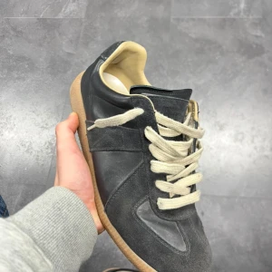 Maison Margiela GAT sneakers i svart skinn - Maison Margiela GAT sneakers i svart skinn med vita snören och beige sula. Klassisk och stilren design med rund tå och platt sula. Perfekta för dig som gillar minimalistiska och tidlösa sneakers. Säljer skorna på grund av att jag har haft dem ett tag, och söker nya alternativ. Skorna är i  väldigt gott skick och har fortfarande mycket kvar att ge. Är även öppen för byten och priset kan diskuteras vid snabb affär! , digitalt kvitto finns!