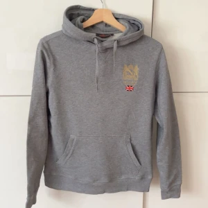 Morris hoodie - Grå hoodie från Morris med broderad gulddetalj och brittisk flagga på bröstet. Tröjan har huva med dragsko och en stor magficka framtill. Perfekt för en avslappnad stil.