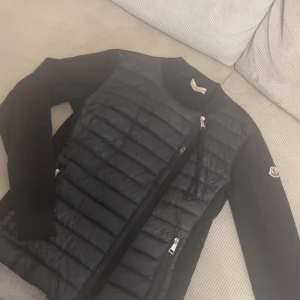 Svart cardigan från Moncler - Säljer en svart pufferjacka från Moncler med quiltad front och släta ärmar. Jackan har dragkedja framtill och klassisk Moncler-logga på ärmen. Perfekt för dig som vill ha en stilren och exklusiv look.