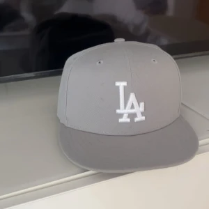 LA keps - Säljer en stilren grå keps från New Era med LA-logga broderad framtill i vitt. Klassisk 59FIFTY-modell med rak skärm och broderade ventilationshål. Perfekt för dig som gillar sportig streetstyle.