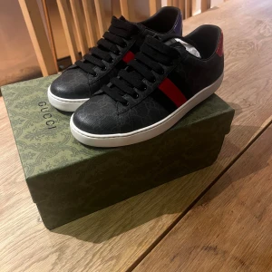 Gucci ace skor svarta - Säljer ett par svarta Gucci sneakers med diskret GG-mönster och röda ränder på sidorna. Skorna har vita platta sulor och svart snörning. Klassisk och stilren design med exklusiv känsla.