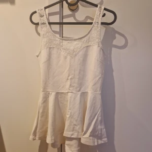 Vit ärmlös klänning med spetsdetaljer - Söt vit klänning med ärmlös design och spetsdetaljer upptill. Klänningen har en utställd kjol och rundad halsringning, perfekt för sommarens varma dagar.