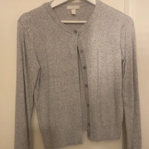 Ljusgrå kofta från H&M - Säljer en ljusgrå kofta från H&M Basics. Koftan har rund halsringning, långa ärmar och knäppning framtill med små knappar. Perfekt att slänga över en topp eller klänning för en enkel och stilren look.