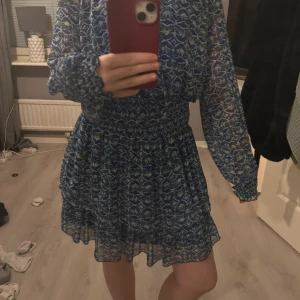 Blåvit mönstrad klänning från Zara - Supersöt blå och vit mönstrad klänning från Zara med långa ärmar och smock i midjan. Klänningen har v-ringning med knytband och volangdetaljer vid ärmslut och kjol. Perfekt för vår och sommar!