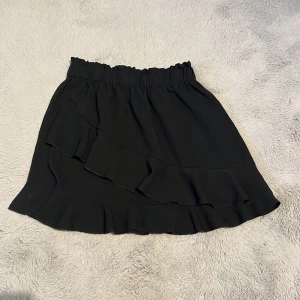 Svart kortkjol med volang - Snygg svart kortkjol med volangdetaljer. Perfekt för att ge en stilren look med en touch av elegans. Den elastiska midjan gör den bekväm att bära hela dagen.