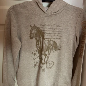 Beige hoodie från U.S. Polo Assn. - Säljer en beige hoodie från U.S. Polo Assn. med ett glittrigt hästmotiv och text på framsidan. Tröjan har långa ärmar och en bekväm huva. Perfekt för en avslappnad stil. (aldrig använt)