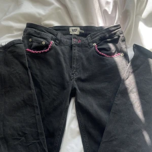 Svarta jeans med leoparddetaljer och rosa sömmar - Coola svarta jeans från lager 157 med unika leopardmönstrade detaljer på bakfickorna och rosa sömmar. Byxorna har en hjärtformad detalj på ena fickan och är i storlek M med kort längd. Perfekta för att ge din outfit en edgy touch!