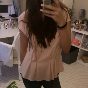 Rosa blus med volangdetalj - Säljer en stilren rosa blus med volangdetalj i nederkant. Blusen har korta ärmar och en lös passform som ger en avslappnad look. Perfekt för både vardag och speciella tillfällen.
