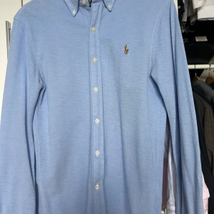 Blå skjorta från Ralph Lauren - Snygg blå skjorta från Ralph Lauren med klassisk krage och broderad logotyp på bröstet. Skjortan har långa ärmar och knappar framtill. Perfekt för en stilren look. Oxford knitted slimfit modell. Använd sparsamt, men tvätthål vid ryggvecket/under tag i nacken som endast syns på insidan.