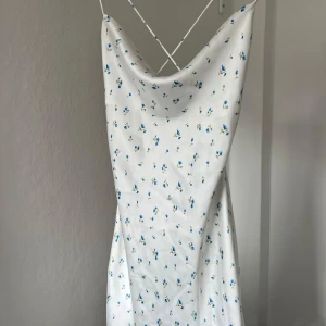 Vit blommig klänning med öppen rygg - Säljer en söt vit klänning med små blå blommor och smala axelband. Klänningen har en öppen rygg med snörning och är perfekt för sommarens varma dagar. Den är ärmlös och har en lätt och luftig känsla. Finns inte att köpa längre🩷