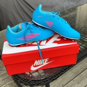 Blå fotbollsskor från Adidas - Snygga blå fotbollsskor från Adidas med rosa detaljer och snörning. Skorna har en robust sula med dobbar för bra grepp på planen. Perfekta för fotbollsträning och matcher.