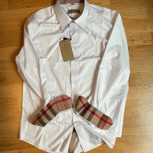 Vit skjorta från Burberry - Säljer en stilren vit skjorta från Burberry med klassiska rutor på insidan av kragen och ärmsluten. Den är helt oanvänd. Hör av dig vid funderingar! (Säljer åt en vän!)