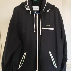 Säljer en svart vindjacka från Lacoste med dragkedja och huva. Jackan har vita detaljer och en broderad logotyp på bröstet. Praktiska fickor med dragkedja och snörning vid huvan för justerbar passform.    !Pris ej hugget i sten!