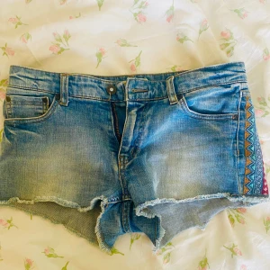 Jeansshorts med broderade detaljer - Snygga jeansshorts med fransiga kanter och färgglada broderade mönster på sidorna. Perfekta för en avslappnad sommarlook. De har en klassisk femficksdesign och knappgylf.
