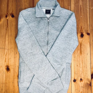 Grå tröja med dragkedja från Superdry - Säljer en stilren grå tröja från Superdry med dragkedja och hög krage. Tröjan har långa ärmar och ribbade muddar vid ärmslut och nederkant. Perfekt för en avslappnad look.