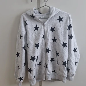 Grå hoodie med stjärnor  - Säljer en grå hoodie från Lindex med svarta stjärnor över hela. Tröjan har dragkedja framtill och en mysig huva. Storlek 170. Använd ett få antal gånger 