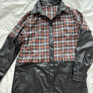 Rutig jacka med skinnärmar från Ganni - Unik jacka från Ganni med rutig design i orange och grått samt svarta skinnärmar. Jackan har knappar framtill och en klassisk krage. Perfekt för en trendig look!