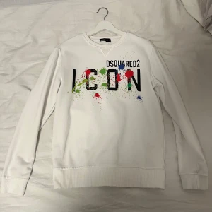 Vit tröja från Dsquared2 - Säljer en vit tröja från Dsquared2 med texten 'ICON' och färgstänk i grönt, rött och blått. Tröjan har långa ärmar och en rund halsringning. Perfekt för en stilren och modern look. Priset sänks vid snabb affär.