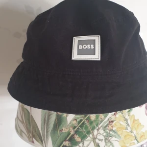 Svart hatt från BOSS - Snygg svart hatt från BOSS med en stilren design. Hatten har en fyrkantig logotyp framtill och är perfekt för att ge en cool touch till din outfit. Tillverkad i ett bekvämt material som passar bra för olika tillfällen.