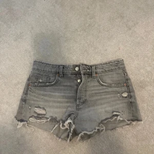 Grå jeansshorts från Zara - Säljer ett par grå jeansshorts från Zara med slitna detaljer och fransar. Shortsen har en låg midja och klassisk femficksdesign. Perfekta för en avslappnad stil.
