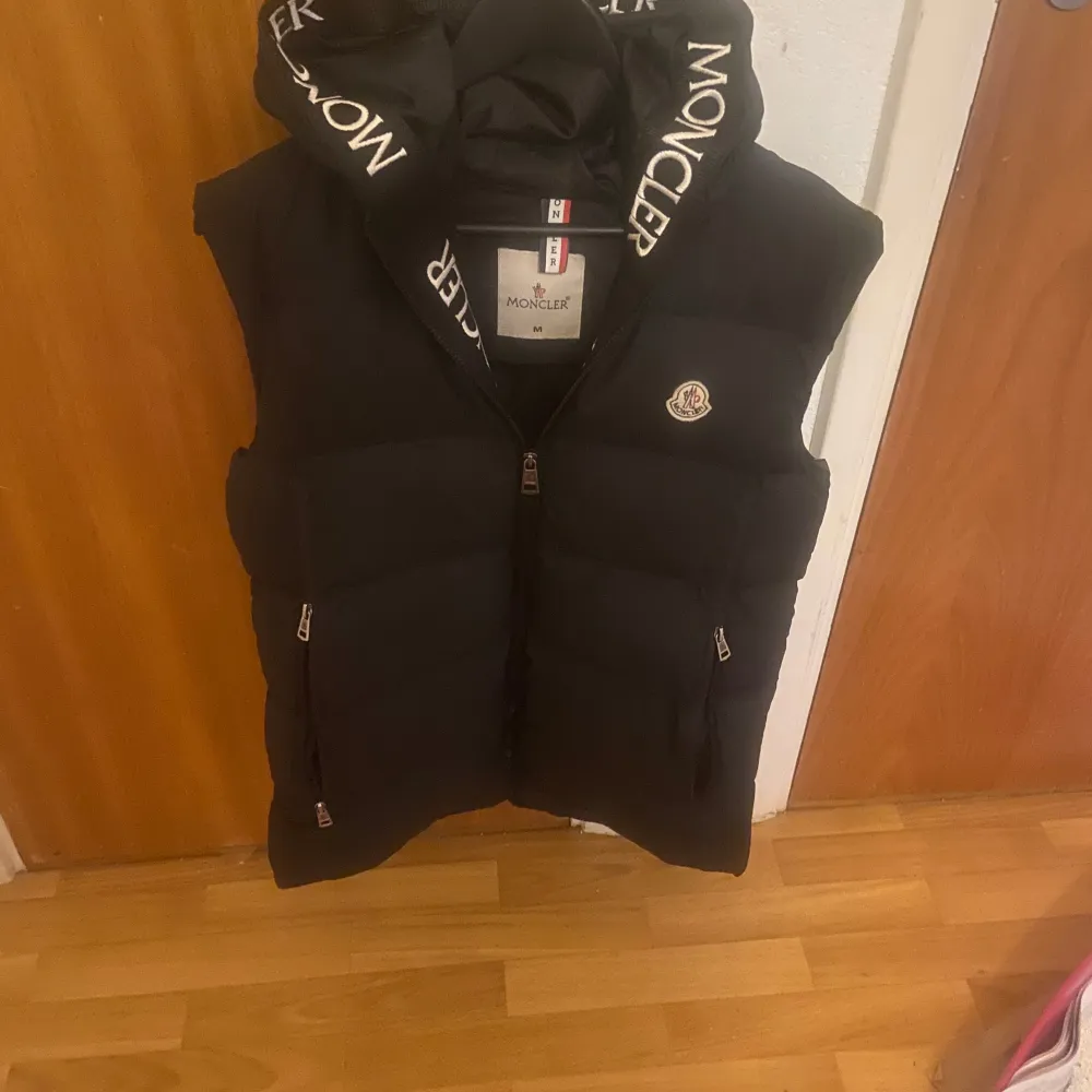 Snygg svart dunväst från Moncler med dragkedja och huva. Västen har Moncler-logga på bröstet och text på huvan. Perfekt för våren och sommaren när du vill hålla stilen.. Takit.