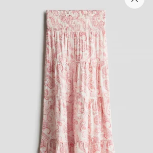 Rosa mönstrad långkjol - Charmig rosa långkjol med ett detaljerat paisleymönster i rött. Kjolen har en luftig passform och är perfekt för varma dagar. Den är tillverkad i ett lätt och bekvämt material som ger en skön känsla.