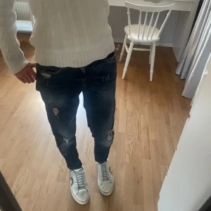 Blå jeans med slitningar - Snygga blå jeans med coola slitningar och en klassisk femficksdesign. De har en normal passform och är perfekta för en avslappnad stil. Jeansen har en knappgylf och är tillverkade i ett robust denimtyg. Tror att modellen på jeansen heter ”Anna”.