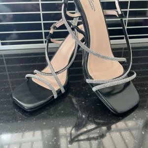 Svarta sandaler med glittriga remmar - Eleganta svarta sandaler med glittriga remmar och transparent klack. De har en fyrkantig tå och justerbara spännen för perfekt passform. Perfekta för en kväll ute eller speciella tillfällen.