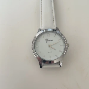Vit klocka från Geneva - Elegant vit klocka från Geneva med ett stilrent quartz-urverk. Klockan har en vit urtavla med silverfärgade markeringar och en glittrig kant. Armbandet är gjort av skinn och har en klassisk design.