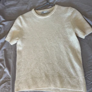 Beige kortärmad tröja från Zara - Säljer en stilren kräm vit tröja från Zara. Den är kortärmad med ribbade kanter vid ärmslut och nederkant. I storlek M