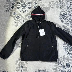 Svart vindjacka från Moncler - Snygg svart vindjacka från Moncler med huva och dragkedja. Jackan har en stilren design med Moncler-logga på ärmen och en randig detalj på huvan. Perfekt för blåsiga dagar.
