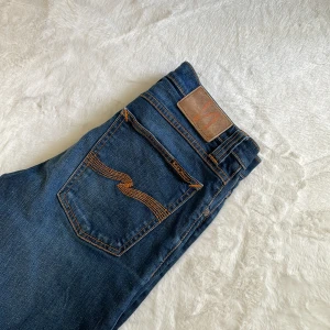Blå jeans från Nudie Jeans - Snygga blå jeans från Nudie Jeans med klassisk femficksdesign och orange kontrastsömmar. Byxorna har en knappgylf och en läderpatch med logotyp baktill. Perfekta för en stilren look.