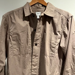 Beige overshirt från H&M - Snygg beige overshirt från H&M med klassisk krage och knappar framtill. Skjortan har två bröstfickor och är perfekt för en stilren look.