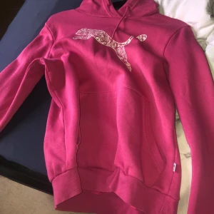 Rosa hoodie från Puma - Säljer en snygg rosa hoodie från Puma med det klassiska logotyptrycket på bröstet. Tröjan har en stor ficka framtill och en bekväm huva. Perfekt för en avslappnad stil.
