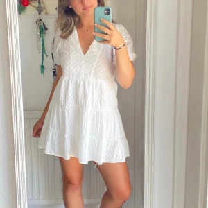 Vit klänning från Zara - Säljer en söt vit klänning från Zara med v-ringning och korta ärmar. Klänningen har ett fint broderat mönster och är perfekt för sommardagar. Den är kort och luftig, vilket gör den bekväm att bära.