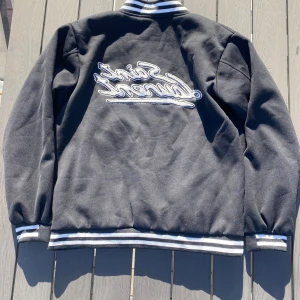 Svart bomberjacka från Saint Laurent - Snygg svart bomberjacka från Saint Laurent med vita detaljer. Jackan har knappar framtill och ribbade muddar vid ärmslut och nederkant. Den har också två vita fickor och en broderad logga på ryggen. Perfekt för en stilren look!