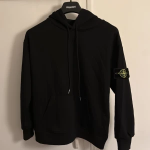 Svart hoodie från Stone Island - Säljer en stilren svart hoodie från Stone Island med klassisk logga på ärmen. Tröjan har en bekväm passform med långa ärmar och en praktisk magficka. Perfekt för en avslappnad stil.