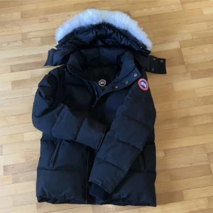 Svart dunjacka från Canada Goose - Säljer en svart dunjacka från Canada Goose med avtagbar huva och vit pälsdetalj. Jackan har en dragkedja och knappar framtill samt en ikonisk logga på ärmen. Perfekt för kalla vinterdagar. Storleken är S men sitter som M.
