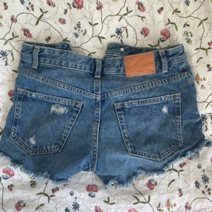 Blå jeansshorts med fransar - Snygga blå jeansshorts med slitna detaljer och fransar vid benen. Perfekta för en avslappnad stil. De har en klassisk femficksdesign och knappgylf. Köpta utomlands❤️
