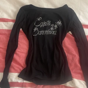 Svart långärmad topp med strassdetaljer - Snygg svart långärmad topp med glittrande strassdetaljer som bildar texten 'Capita en Damelona'. Perfekt för att ge en touch av glamour till din outfit. Passar bra till både jeans och kjol.